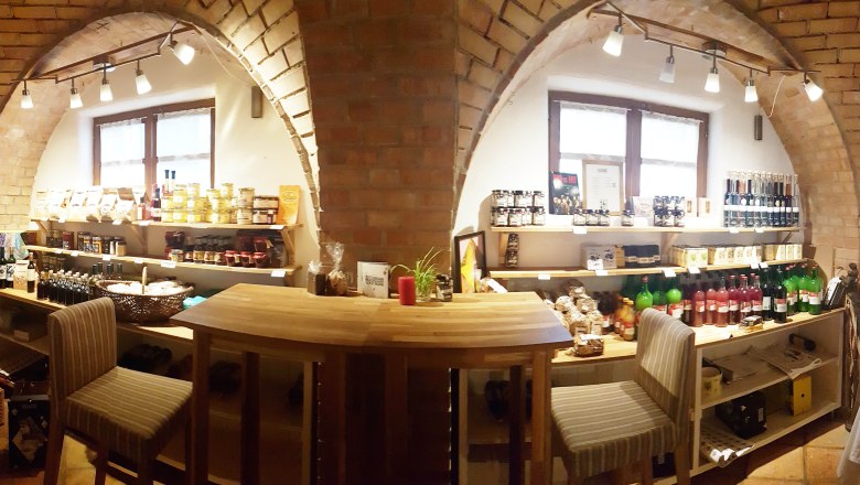 Greißlerei, © Zuschmann-Schöfmann Innenansicht eines kleinen Ladens mit Regalen voller Lebensmittel und Getränken, Holztisch und Stühlen.