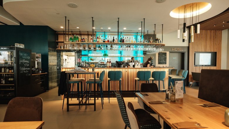 freilich Barbereich, © Michael Reidinger Moderner Barbereich mit Holztischen und blauen Barhockern, beleuchtet durch hängende Lampen.