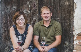 Barbara und Christoph Pletzer, © Anna Zora Ein Mann und eine Frau sitzen vor einer Holzwand und halten Weingläser in den Händen.