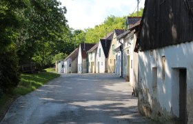 Kellergasse Altlichtenwarth, © Weinviertel Tourismus Reihe von Weinkellern in einer ländlichen Straße mit Bäumen im Hintergrund.