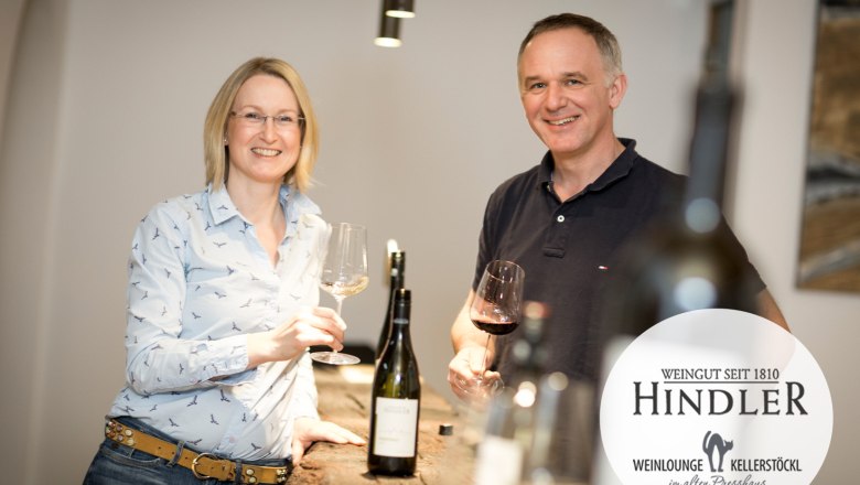 Rosi und Karl Hindler, © Weingut Hindler Ein Winzerpaar steht lächelnd mit Weingläsern in der Hand an einem Tisch im Weingut Hindler.