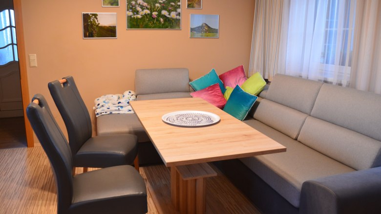 Woh- und Essbereich, © Eva Stuhr Wohn- und Essbereich mit grauem Sofa, Holztisch, bunten Kissen und Wandbildern.