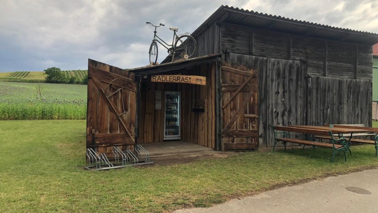 Radlerrast Pottenhofen, © Lukas Cermak Eine rustikale Holzhütte mit der Aufschrift 'Radlerrast' und einem Fahrrad auf dem Dach, umgeben von Feldern und einer Bank.