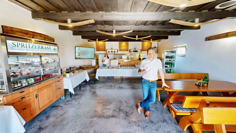 Spritzerrast – Campingstellplatz mit Jause und Panoramablick, © Bernhard Raab Ein Mann steht in einem rustikalen Raum mit Holzmöbeln und einem Kühlschrank voller Getränke. Ein Schild mit der Aufschrift 'Spritzerrast' ist sichtbar.