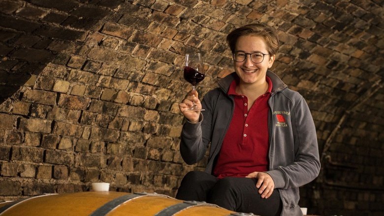 Jungwinzerin, © Paul Breuss Eine Person sitzt auf einem Weinfass in einem Weinkeller und hält ein Glas Rotwein in der Hand.