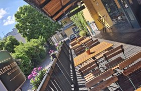 Terrasse Depot, © Altes Depot Mistelbach Eine sonnige Terrasse mit Holztischen und -stühlen, umgeben von Pflanzen und einem Gebäude mit gelber Fassade.