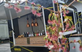 Unterwegs im Foody, © Die zwoa Weinviertla Ein schwarzer Foodtruck mit der Aufschrift 'Genuss Fahrbrik' steht im Freien. Im Vordergrund sind unscharfe Blüten zu sehen.