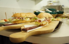 Heurigenjause, © Fam. Zöchmeister Zwei belegte Baguettes auf Holzbrett mit Messer, im Hintergrund weitere Speisen.