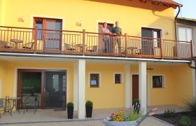 Gästehaus Woditschka, © Fam. Woditschka Zweistöckiges gelbes Gebäude mit Balkon, auf dem zwei Personen stehen. Im Vordergrund eine Schubkarre mit Blumen.