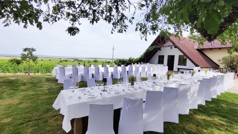 Tafeln im Weinviertel, © Weinviertel Tourismus GmbH Gedeckte Tische im Freien mit weißen Stuhlhussen, umgeben von grüner Landschaft und einem Gebäude mit rotem Dach.
