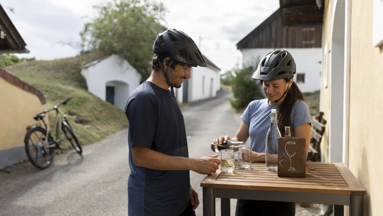 Rest stop in the Öhlbergkellergasse, © Weinviertel Tourismus GmbH / Frühmann Two cyclists with helmets stand at a table in a wine cellar lane and pour themselves drinks.