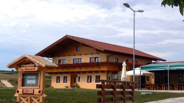 Heurigenlokal Böswirth, © Fam. Böswirth Ein traditionelles Gasthaus mit Holzbalkon und Blumendekoration, daneben ein Schild mit der Aufschrift 'Buschenschank Fam. Böswirth'.