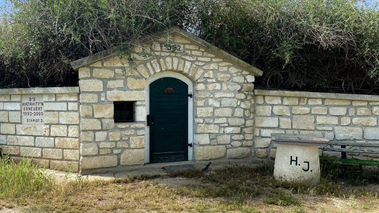 Hiata hut Unterdürnbach, © Weinstraße Weinviertel Stone hut with green gate and inscription, surrounded by bushes and grass.