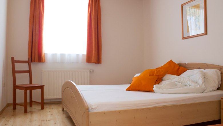 Doppelzimmer, © Fam. Lobner Ein einfach eingerichtetes Doppelzimmer mit Holzbett, orangefarbenen Kissen und Vorhängen, einem Stuhl und einem Bild an der Wand.