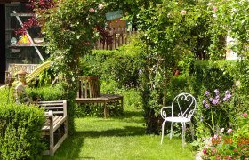 Landhausgarten, © Petra Strobl Ein idyllischer Garten mit Bänken, einem weißen Stuhl und blühenden Pflanzen.