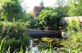Platz am Teich, © Natur im Garten/Martina Liehl-Rainer Ein idyllischer Garten mit Teich, Seerosen und üppiger Vegetation. Im Hintergrund stehen ein Tisch und Stühle auf einer Terrasse.