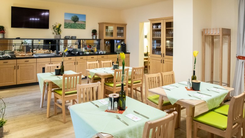 Frühstücksraum, © Nadine Beles Ein gemütlicher Frühstücksraum mit Holzmöbeln, gedeckten Tischen und einem Buffetbereich. An den Wänden hängen Bilder und Pflanzen dekorieren den Raum.