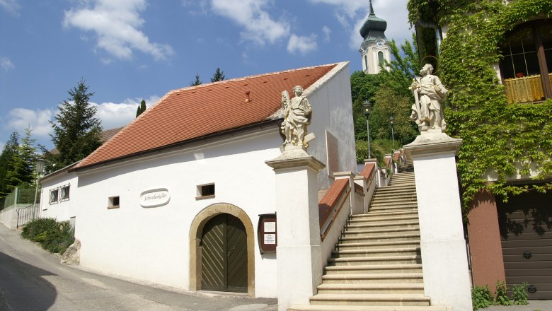 Schwedenkeller, © Stadtgemeinde Mistelbach/Mag. Mark Schönmann Weißes Gebäude mit rotem Dach und Treppe mit Statuen, Kirche im Hintergrund.