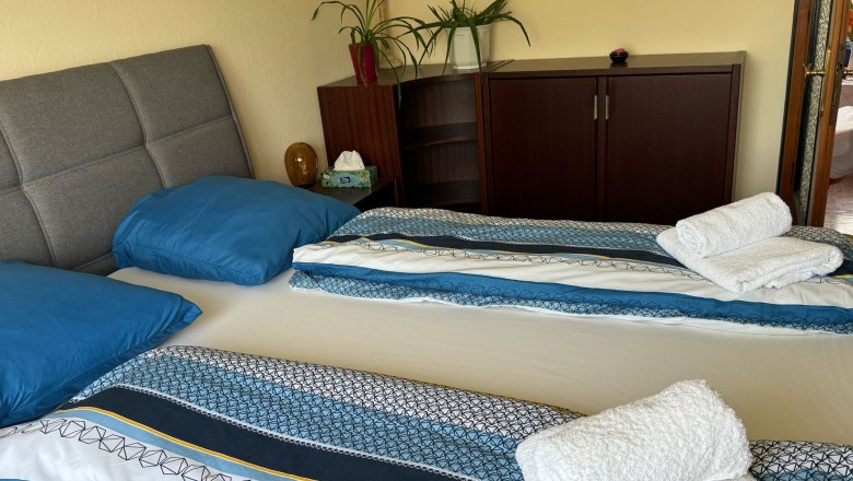 Schlafzimmer, © Dana Perl Ein gemütliches Schlafzimmer mit einem Doppelbett, blauer Bettwäsche und gefalteten Handtüchern. Im Hintergrund ein Schrank mit Pflanzen.