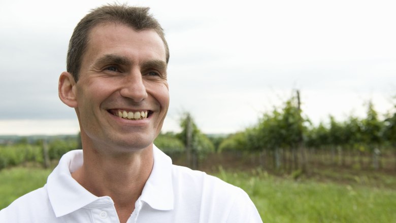 Winzer, © Michael Himml Ein lächelnder Mann in einem weißen Polohemd steht auf einem Feld mit Bäumen im Hintergrund.