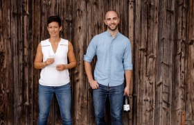 Weingut Jatschka, © Astrid Bartl Zwei Personen stehen vor einer Holzwand, eine Frau mit Weinglas und ein Mann mit Weinflasche.