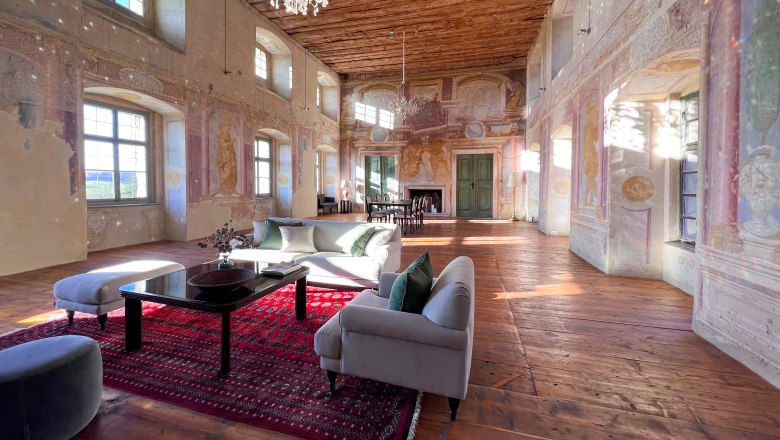 Festsaal, © Schloss Haggenberg Ein historischer Festsaal mit Holzboden, bemalten Wänden und einem Kronleuchter. Moderne Möbel und ein roter Teppich sind im Raum platziert.