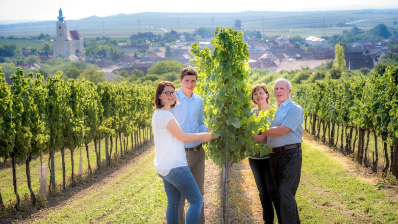 Winzerfamilie Schneider, © Reinhard Podolsky Eine Winzerfamilie steht in einem Weinberg mit einer Kirche im Hintergrund.