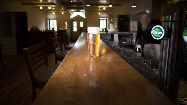 Moravian Embassy, © tonality A glass of beer sits on a long wooden counter in a bar with empty chairs and a person in the background.