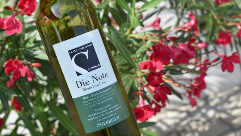 Weinflasche, © Weingut Schober Weinflasche mit Etikett "Die Note" vor blühenden roten Blumen.