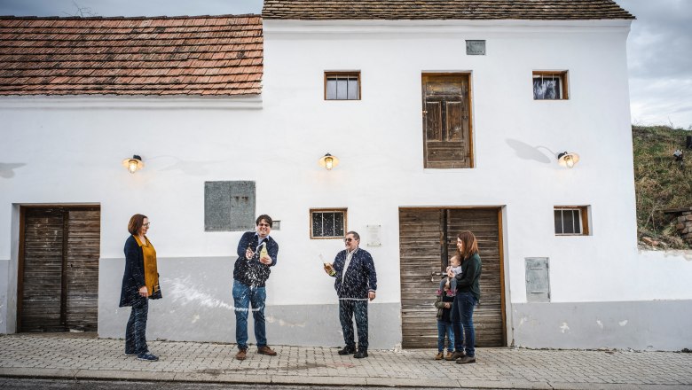 Weinviertler Sektmanufaktur, © Robert Herbst Eine Gruppe von Menschen steht vor einem weißen Gebäude, während eine Person eine Flasche spritzt.