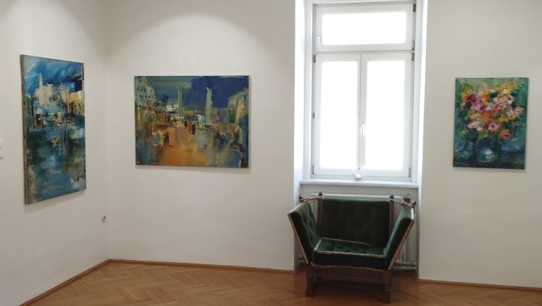 the MERKIMA Gallery, © Doris Hauser Interior view of a gallery with three paintings on the walls and a green armchair in front of a window.