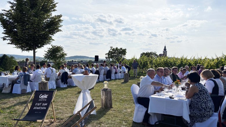 Tafeln im Weinviertel, © Weinviertel Tourismus GmbH Menschen sitzen an gedeckten Tischen im Freien, umgeben von Weinbergen und Bäumen, mit einer Kirche im Hintergrund.