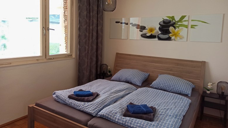 Schlafzimmer, © Multimedia Lauda Schlafzimmer mit Doppelbett, blauer Bettwäsche, Bild mit Steinen und Blumen an der Wand, Fenster und Nachttischlampe.