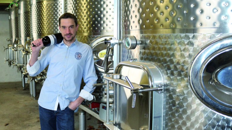 Thomas Mauser, © Martin Mauser Mann in blauer Hemd steht mit Weinflasche vor Edelstahltanks in einer Weinkellerei.