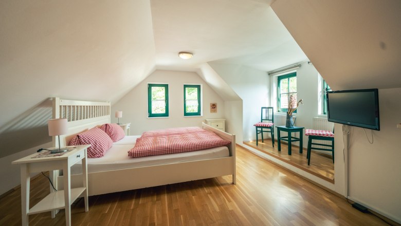 Winery Küssler, © Winzerhof Küssler Bright bedroom with sloping ceiling, double bed, wooden floor, two windows, table with chairs and TV on the wall.