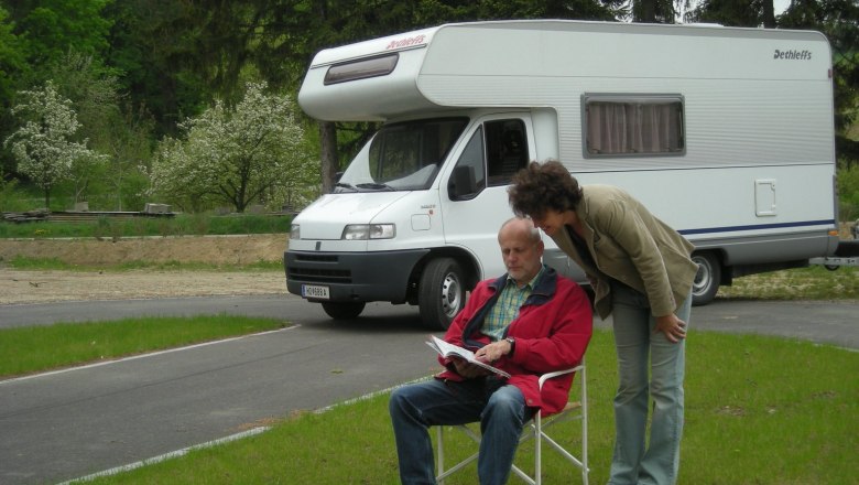 Reisemobil, © S. Satory Ein Mann sitzt auf einem Campingstuhl vor einem Wohnmobil, während eine Frau über seine Schulter schaut. Im Hintergrund sind Bäume und Wiese zu sehen.