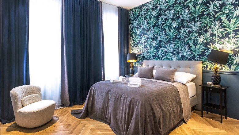 Doppelzimmer, © Astrid Bartl Modernes Doppelzimmer mit grauem Bett, tropischer Tapete und dunklen Vorhängen.