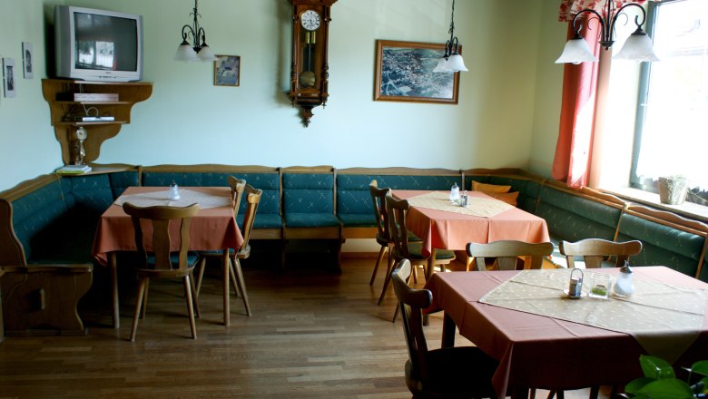 Frühstücksraum, © Privatzimmer Würger Ein traditionell eingerichteter Frühstücksraum mit Holzmöbeln, Tischen mit orangefarbenen Tischdecken, einer Wanduhr und einem Fernseher.