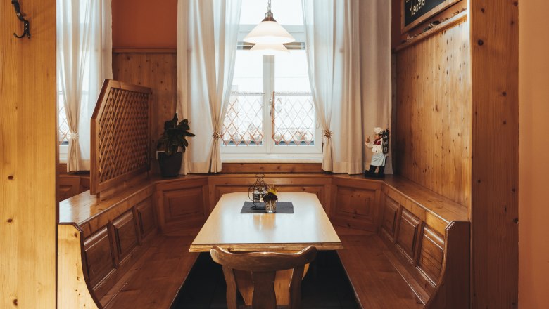 Viel Holz schafft heimeliges Ambiente, © Niederösterreich Werbung/David Schreiber Gemütliche Holzeckbank mit Tisch in einem rustikalen Raum, dekoriert mit Vorhängen und einer kleinen Figur.