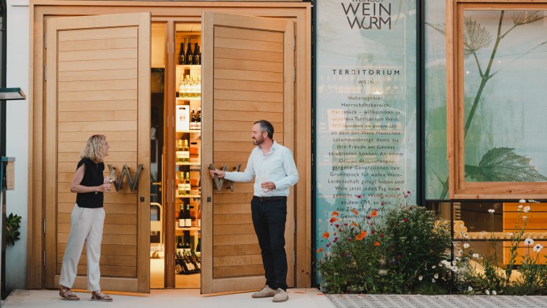 Weingut Weinwurm, © Michael Reidinger Zwei Personen stehen vor einem Weingut mit geöffneten Holztüren, die Flaschen im Inneren zeigen.