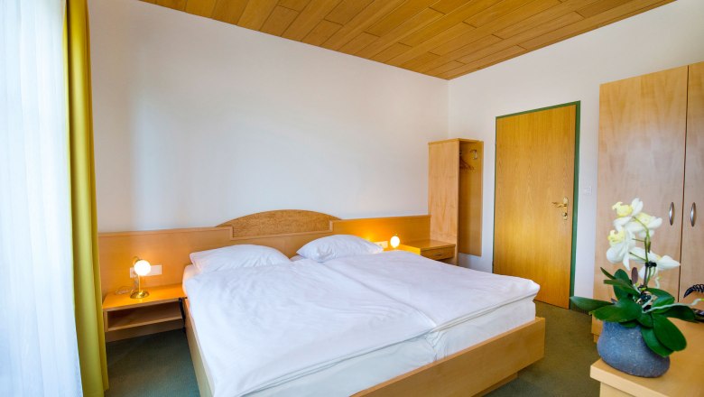 Zimmeransicht, © Hotel Stich Ein einfach eingerichtetes Hotelzimmer mit Doppelbett, Holzdecke, Schrank und Nachttischlampen.