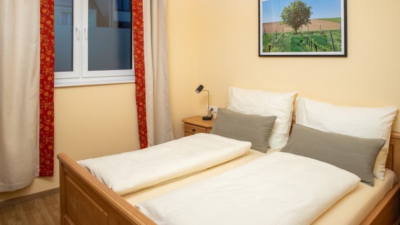 Doppelzimmer, © Nadine Beles Ein gemütliches Doppelzimmer mit einem Doppelbett, einem Fenster mit Vorhängen und einem Landschaftsbild an der Wand.