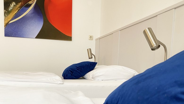 Gästezimmer, © Stadthotel Hollabrunn Ein Gästezimmer mit einem Bett, blauen Kissen, Leselampen und einem abstrakten Gemälde an der Wand.