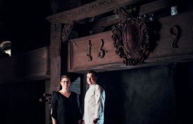 Wirtsleute: Bernadette und Samuel Pope, © Niederösterreich Werbung/Julius Hirtzberger Zwei Personen stehen vor einem alten Holzbalken mit Schnitzereien und der Jahreszahl 1692.