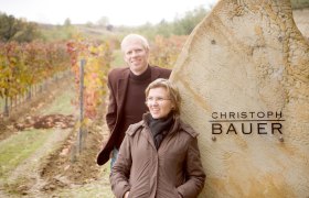 Herbstzeit, © Astrid Bartl Zwei Personen stehen lächelnd vor einem Weinberg neben einem Stein mit der Aufschrift 'Christoph Bauer'.