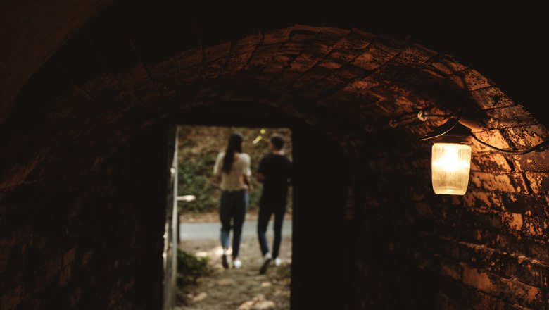 Wine cellar lane Schindergasse, © Weinviertel Tourismus GmbH / Michael Reidinger View from a dark cellar vault of two people outdoors, illuminated by a lamp.