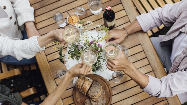 Weinverkostung und Jause, © Weinviertel Tourismus GmbH / Sophie Menegaldo Drei Personen stoßen mit Weingläsern an einem Holztisch an, auf dem Brot, Wasser und Blumen stehen.