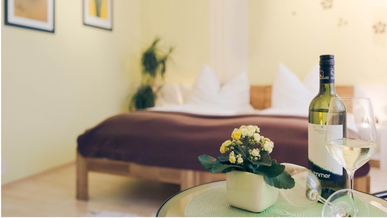 Appartment & Gästezimmer "Vino Verde", © Fam. Willmann Ein gemütliches Schlafzimmer mit einem Bett im Hintergrund und einem Tisch mit einer Weinflasche und einem Glas im Vordergrund.