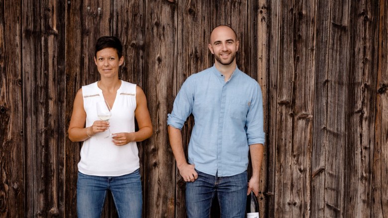 Weingut Jatschka, © Astrid Bartl Zwei Personen stehen vor einer Holzwand, eine Frau hält ein Weinglas, ein Mann eine Weinflasche.