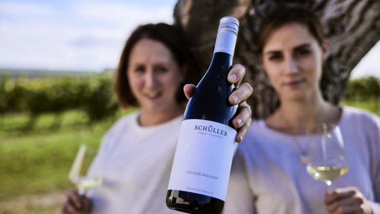 Weingut Schüller, © Stefan Joham Zwei Personen halten eine Flasche Schüller Weißburgunder und Weingläser vor einem Baum in einem Weinberg.